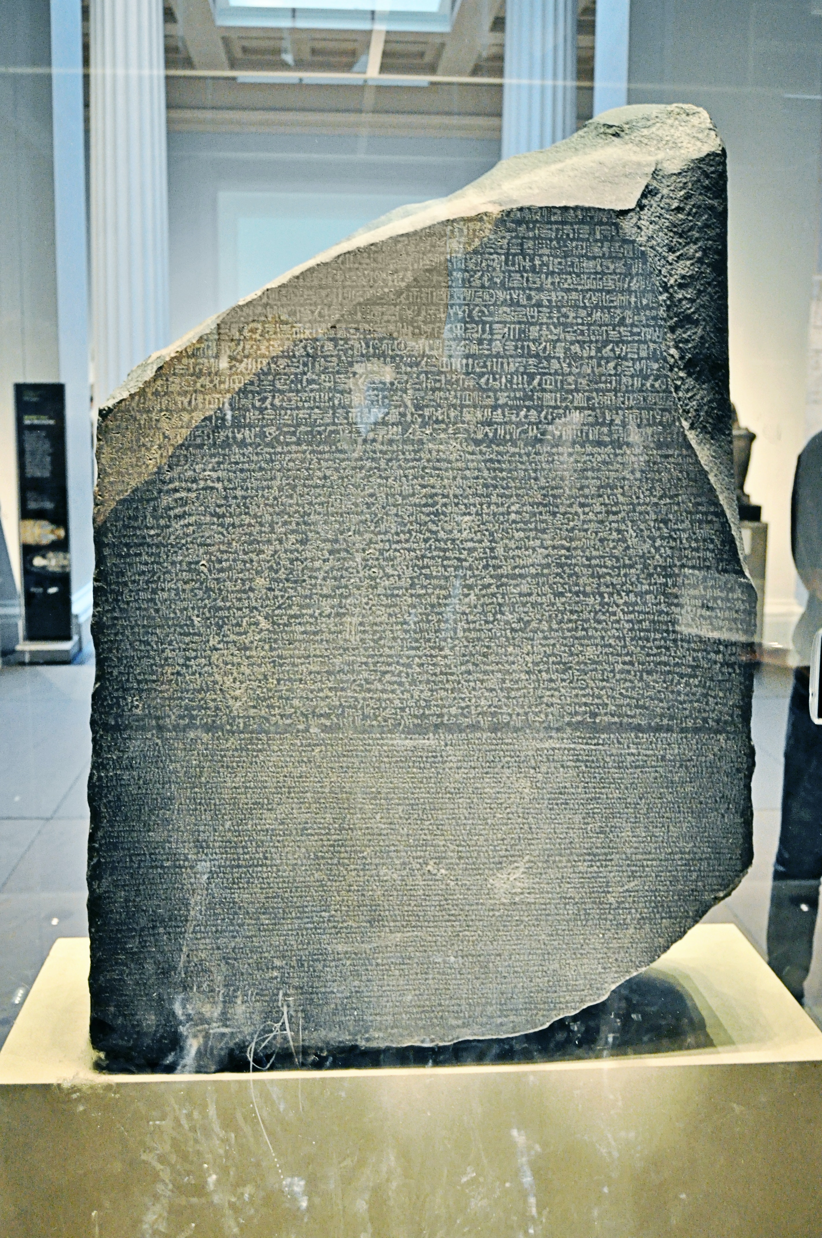 The Rosetta Stone, a slab of stone covered in small inscriptions, stands behind glass casing. The top of the stone is jagged and diagonal where the rest of the tablet has been lost.