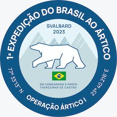 Círculo azul onde se lê: 1a expedição do Brasil ao Ártico, Operação Ártico I, 77º33'13''N, 23º40.216' E. No centro do círculo, azul claro, vê-se o desenho de montanhas e de um urso polar, com os dizeres Svalbard 2023, e uma bandeira do Brasil